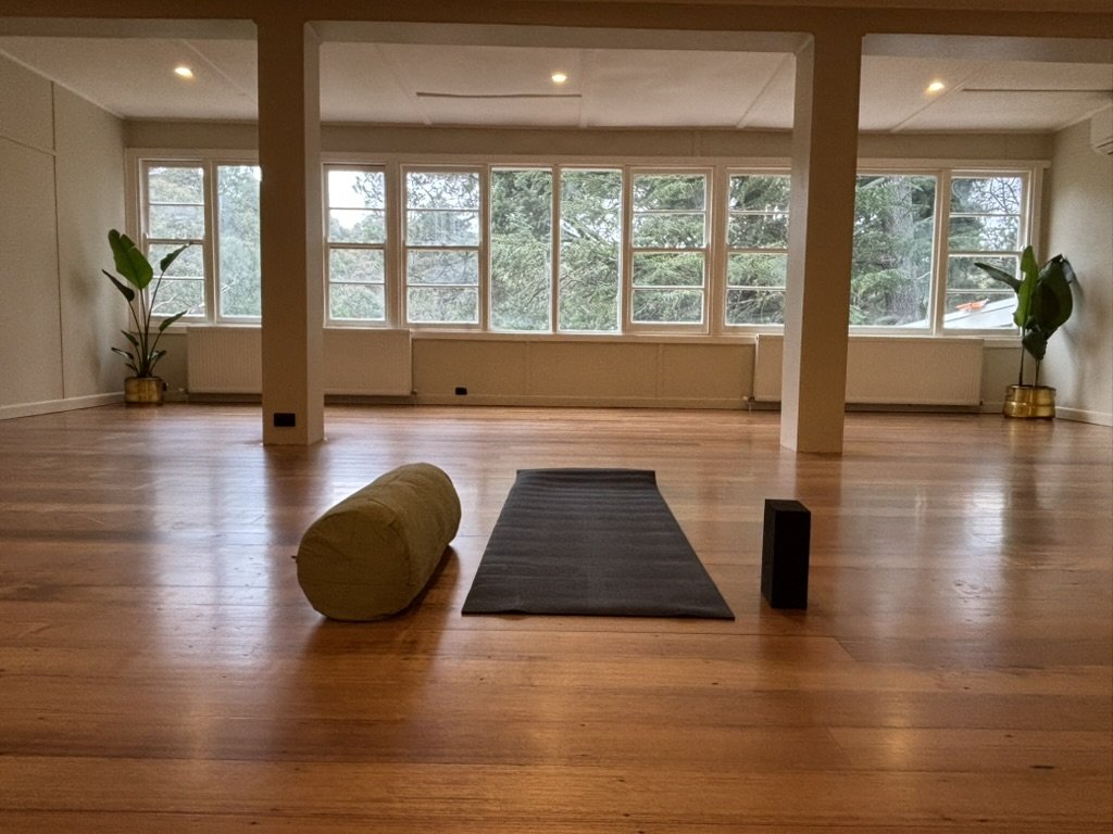 A mat, bolster and block laid out on the hardwood floor of the yoga room, with windows onto the trees